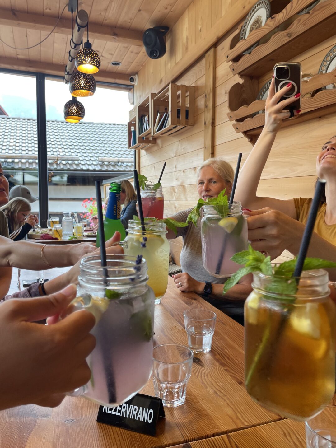 Women clink glasses with mint drinks at a wooden mountain hut café during a hiking stop.