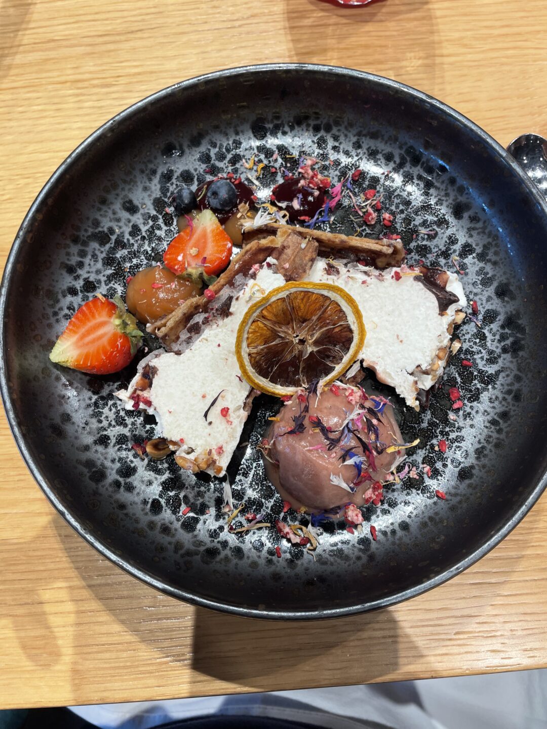 Dessert dish in a black bowl with strawberries, dried orange slice and chocolate, served on a wooden table