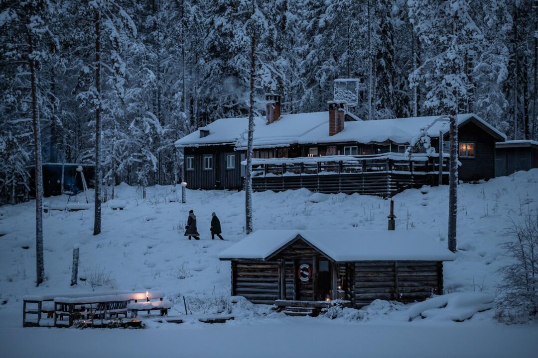 Women walk past wooden cabins in a snowy forest in Sweden, for winter camping in winter conditions.