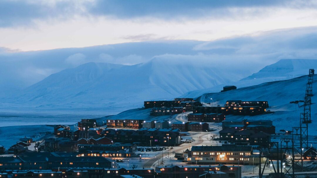 Svalbard settlement at dusk with mountains and illuminated buildings, including harbour shoreline and road lights