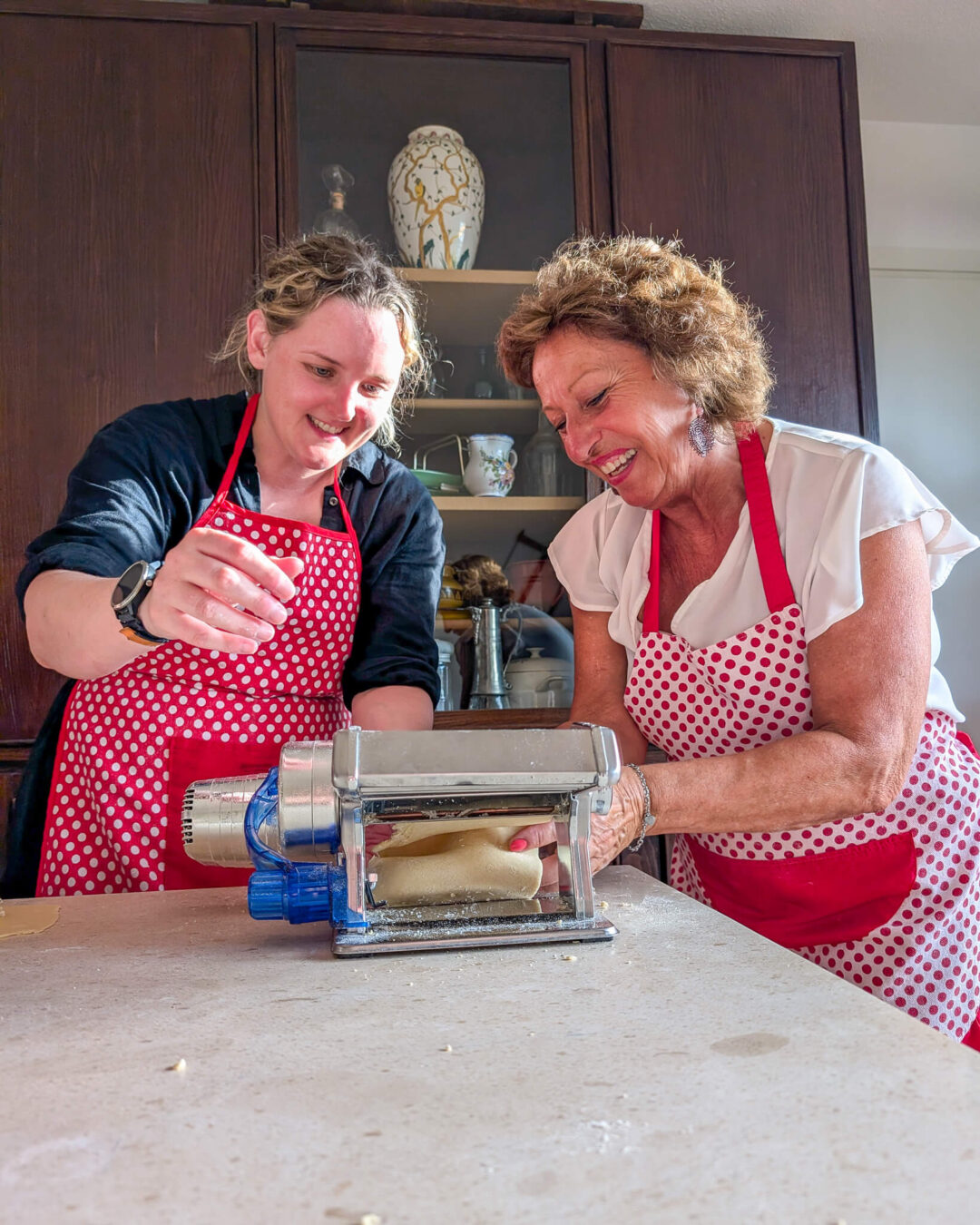 Two women in red polka-dot aprons use a pasta machine in a kitchen.