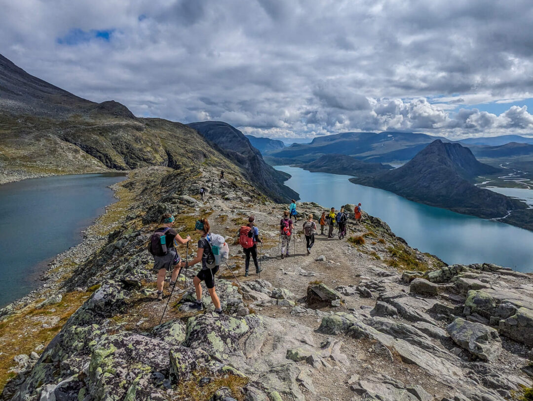Hikers on a rocky ridge during a Norway hiking trek, overlooking blue fjords and mountains under clouds