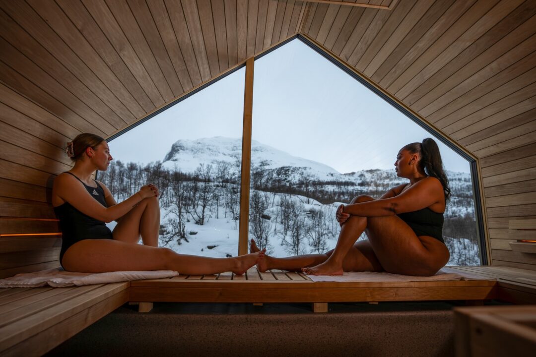 Two women stretching in a wooden sauna with a view of snowy mountains outdoors