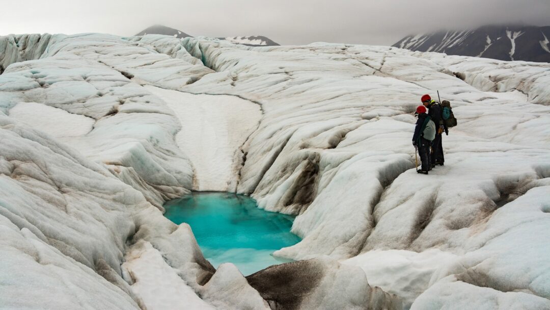 Hiker with backpack trekking across an icy glacier beside turquoise meltwater in the Arctic wilderness