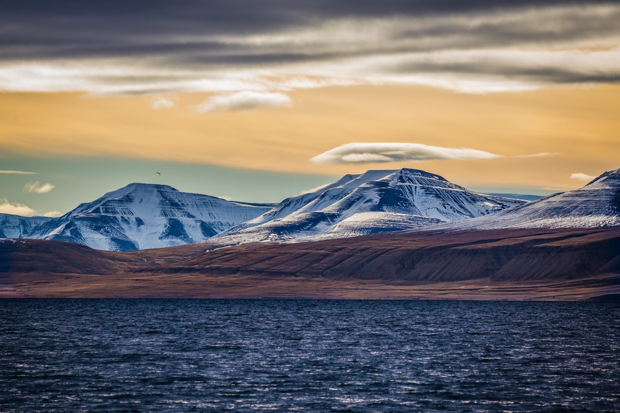 Snowy Arctic mountain peaks over a fjord at dusk, calm sea with dramatic clouds