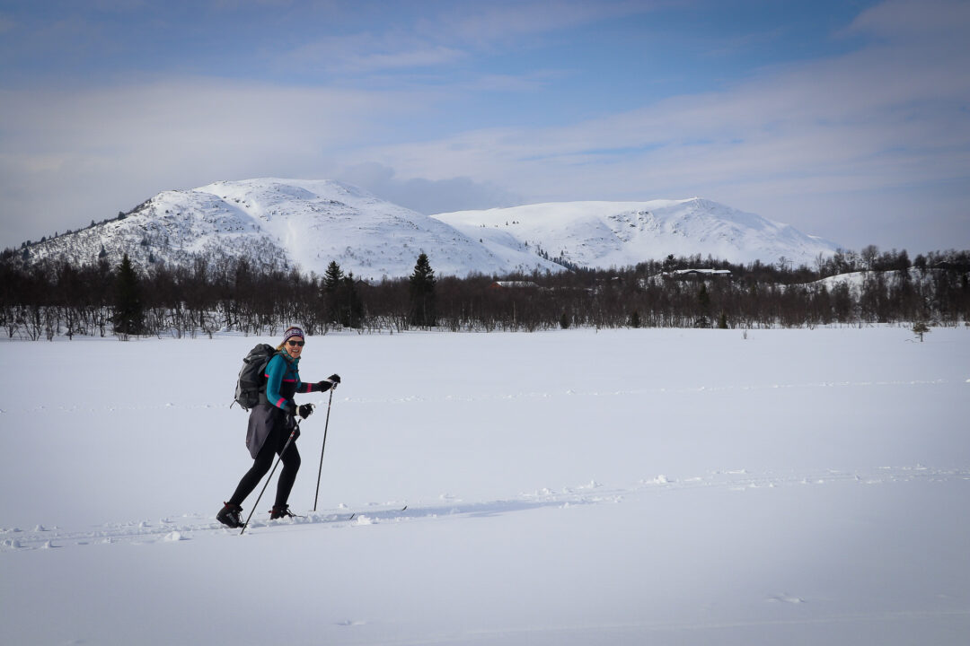 Women cross-country skiing in snowy Norway, gliding across a frozen lake with mountains in the distance.