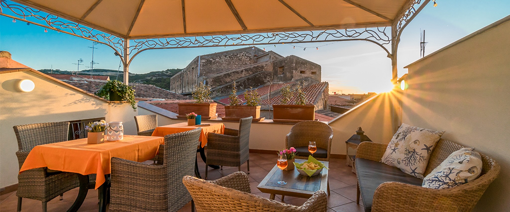 Sunlit outdoor terrace with wicker seating and orange tablecloth, overlooking a rocky hill.