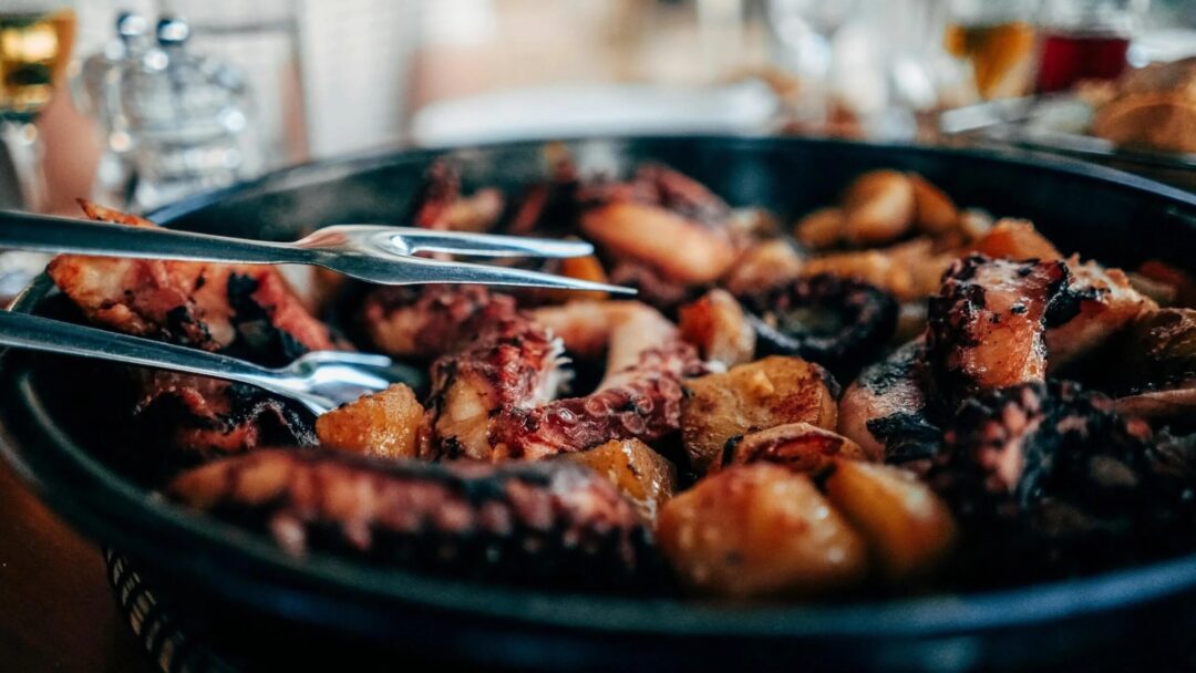 Grilled meat cooking on a barbecue in a black pan, with tongs nearby