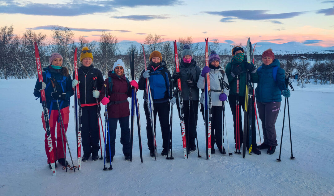 Women cross-country skiers stand together on a snowy Norwegian landscape at dusk with skis and poles