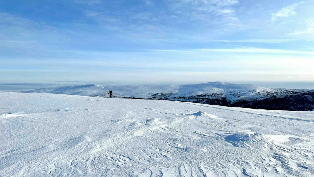Skier crossing a snowy mountain ridge with ski tracks and distant valleys under a blue sky