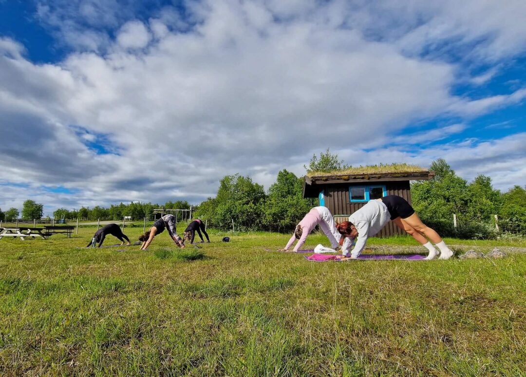 Women doing outdoor stretching on yoga mats in a grassy field beside a stable under blue sky