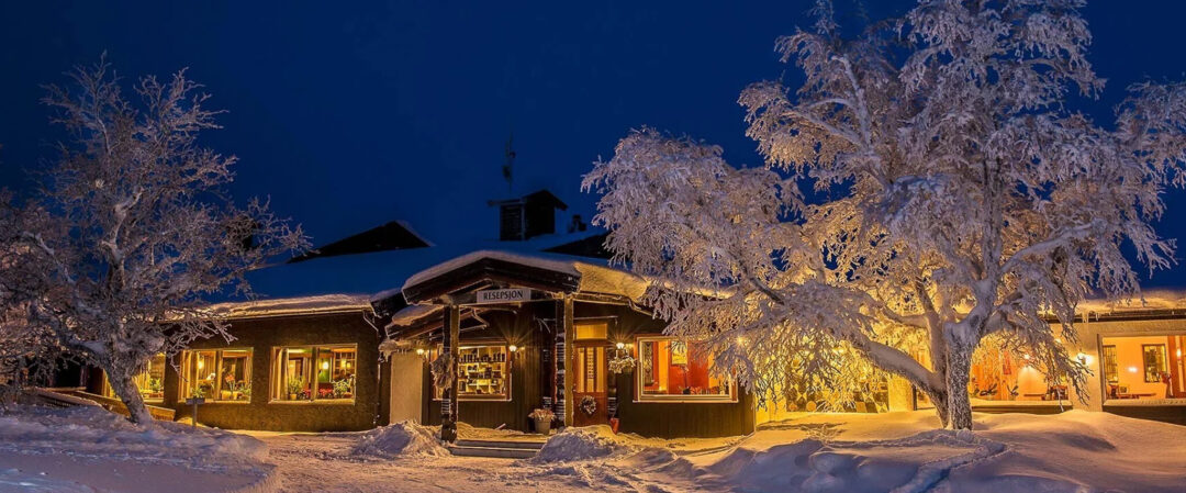 Snowy log cabin at night with warm lights and frosted pine trees