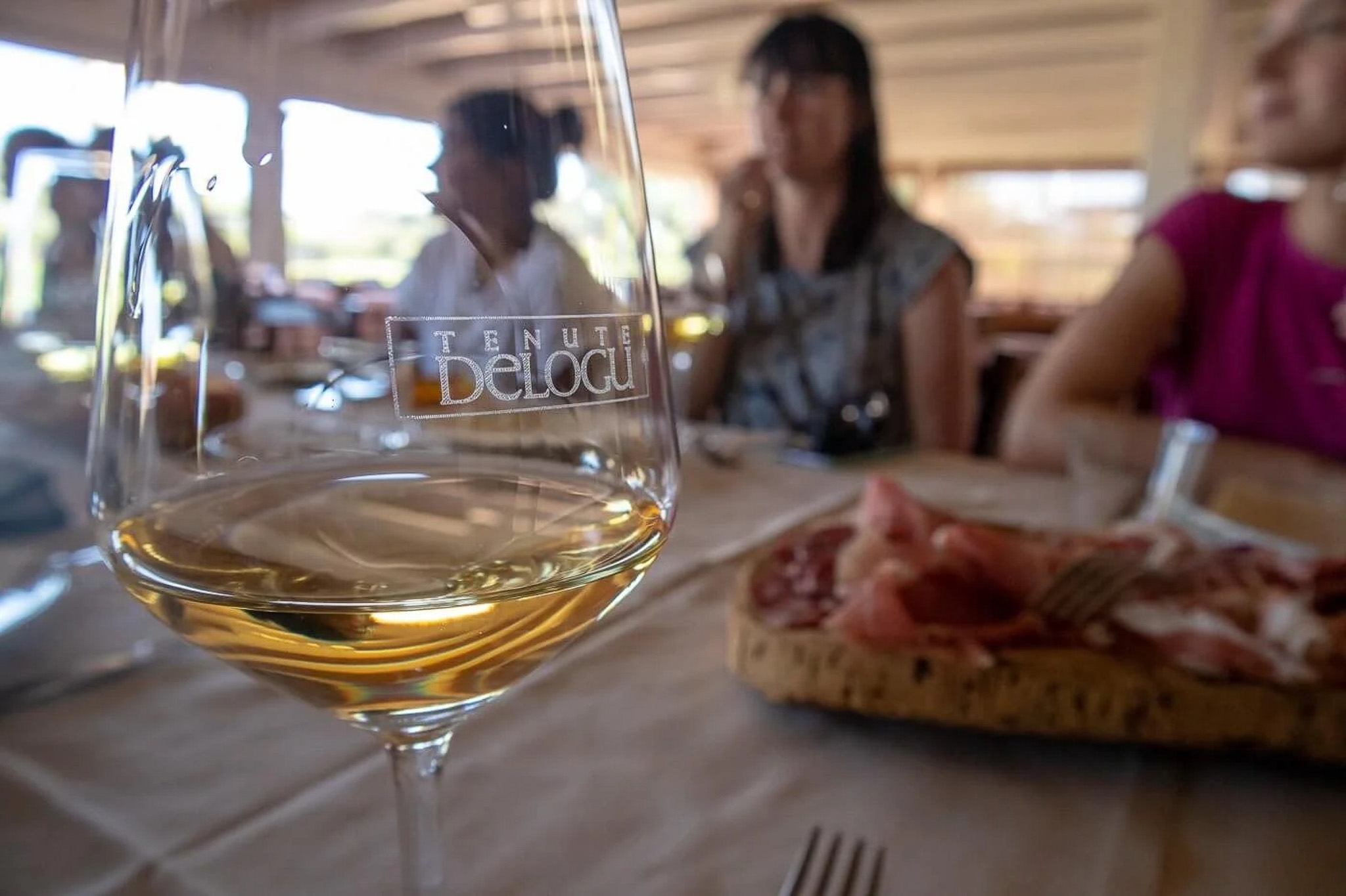 Wine glass labelled “TENUTA Delogu” in foreground, women dining at an outdoor table with food and wine