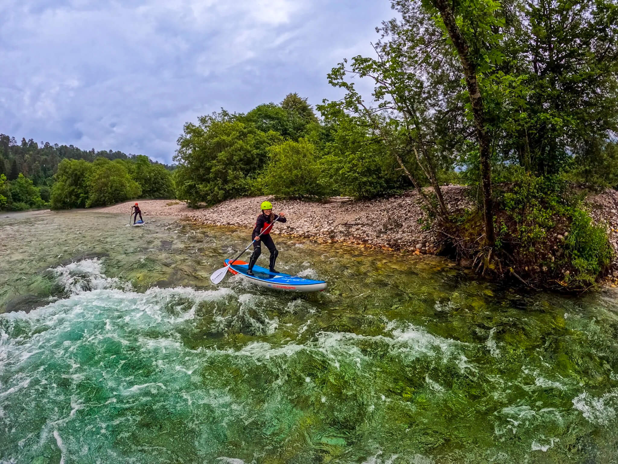 Women stand-up paddleboarding through whitewater rapids on a river with forested banks