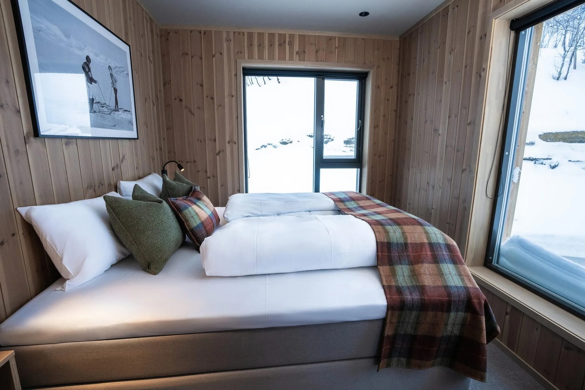 Wooden guest room with bed and plaid blanket, snowy view through large windows in Norway