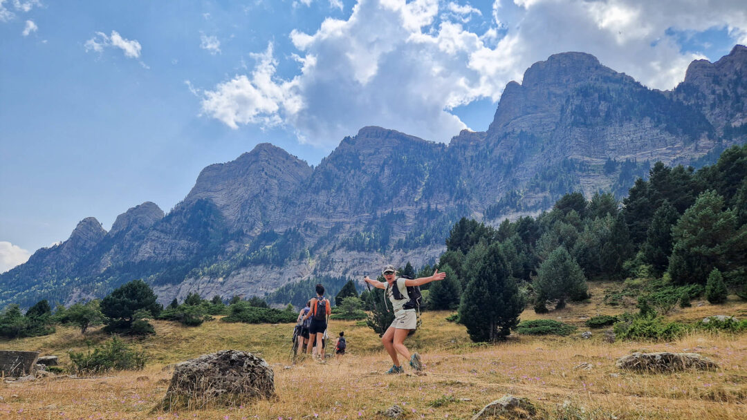 Women hiking on a mountain trail through pine valleys and rocky peaks under a blue sky