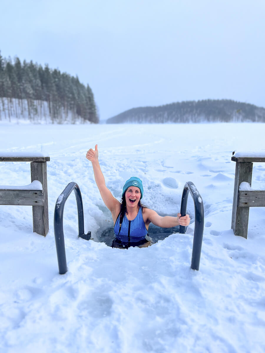 Woman in blue winter gear emerging from a snow plunge, snowy forest landscape under a clear sky