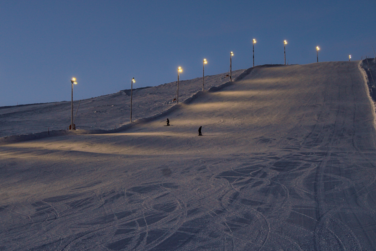 Skiers on a lit downhill ski slope at dusk with visible piste tracks on fresh snow