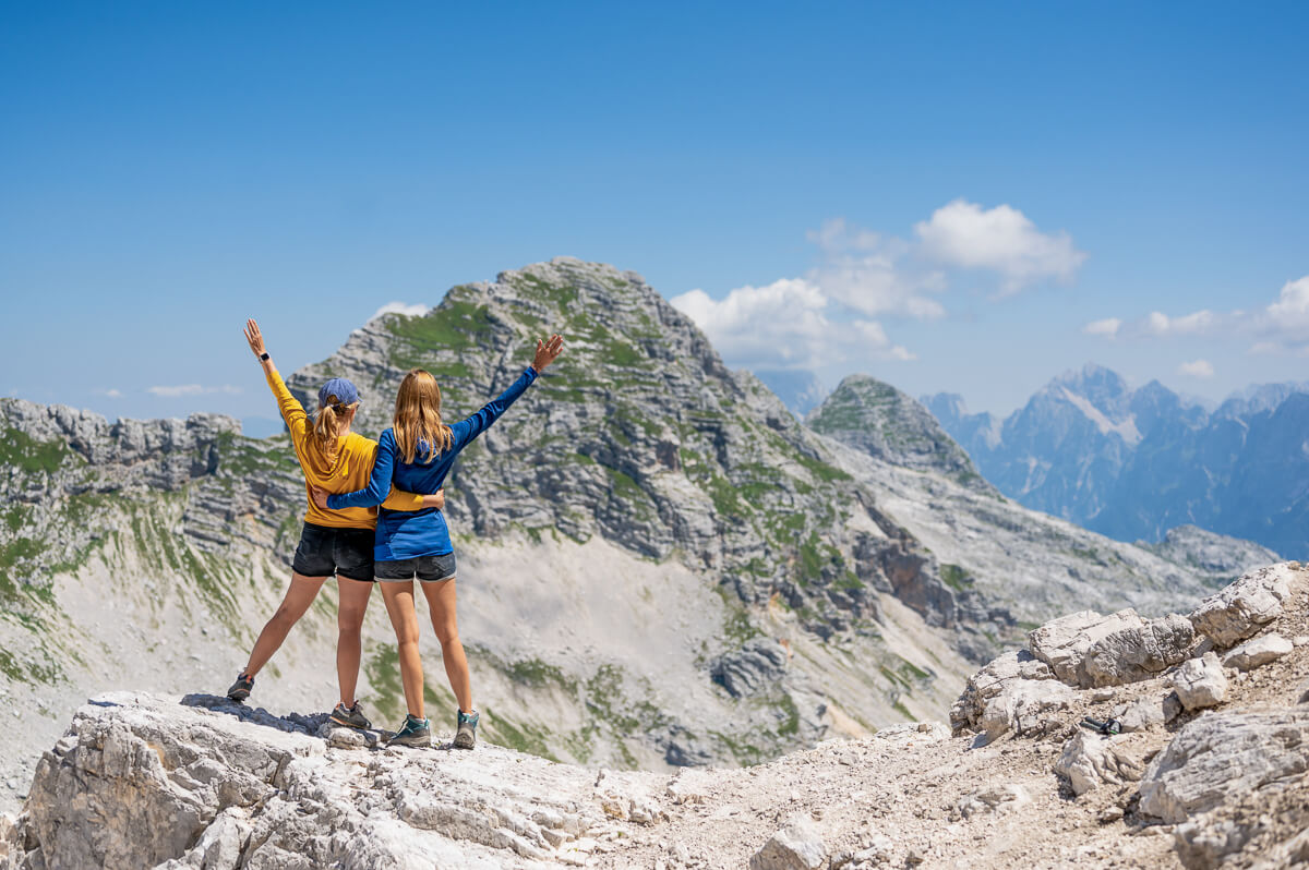 Two women hiking on a rocky ridge with alpine mountains in Slovenia under a blue sky