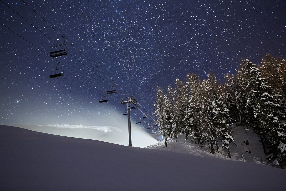 Snowy ski slope at night with chairlift and pine trees under a starry sky