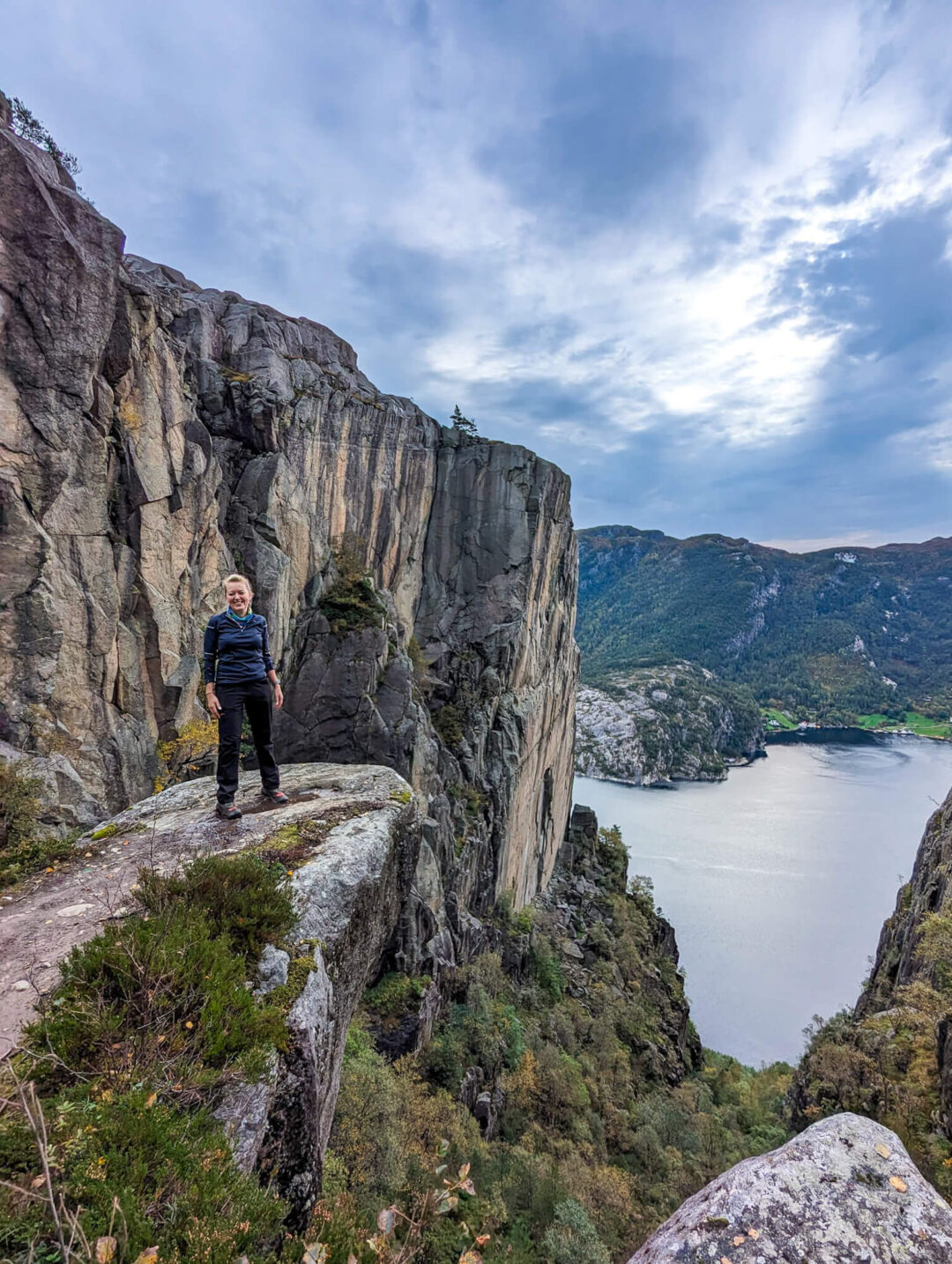 Woman standing on a rocky cliff above a fjord and valley landscape under a cloudy sky