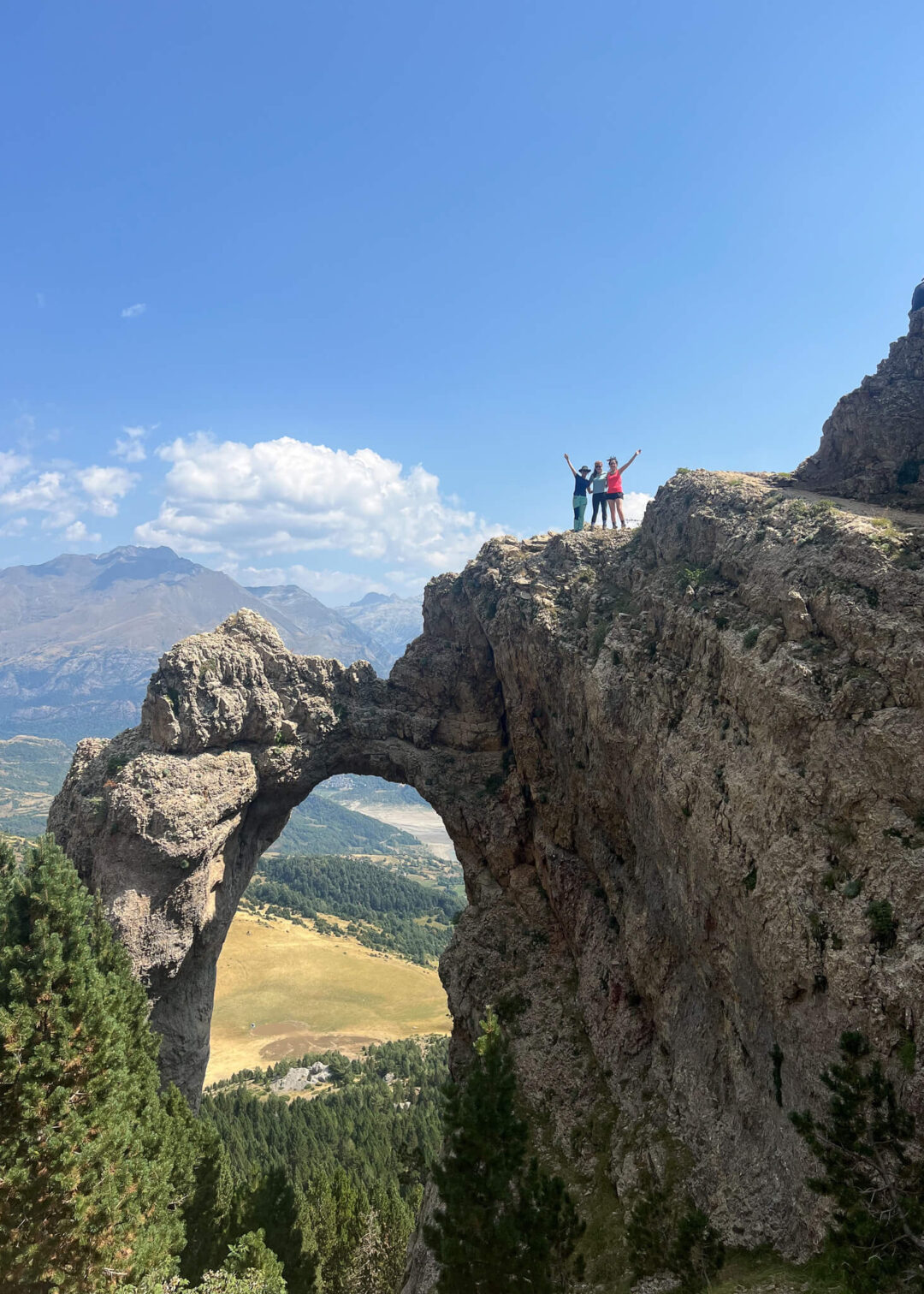 Women hiking on a rocky cliff beside a natural rock arch over a mountain valley viewpoint