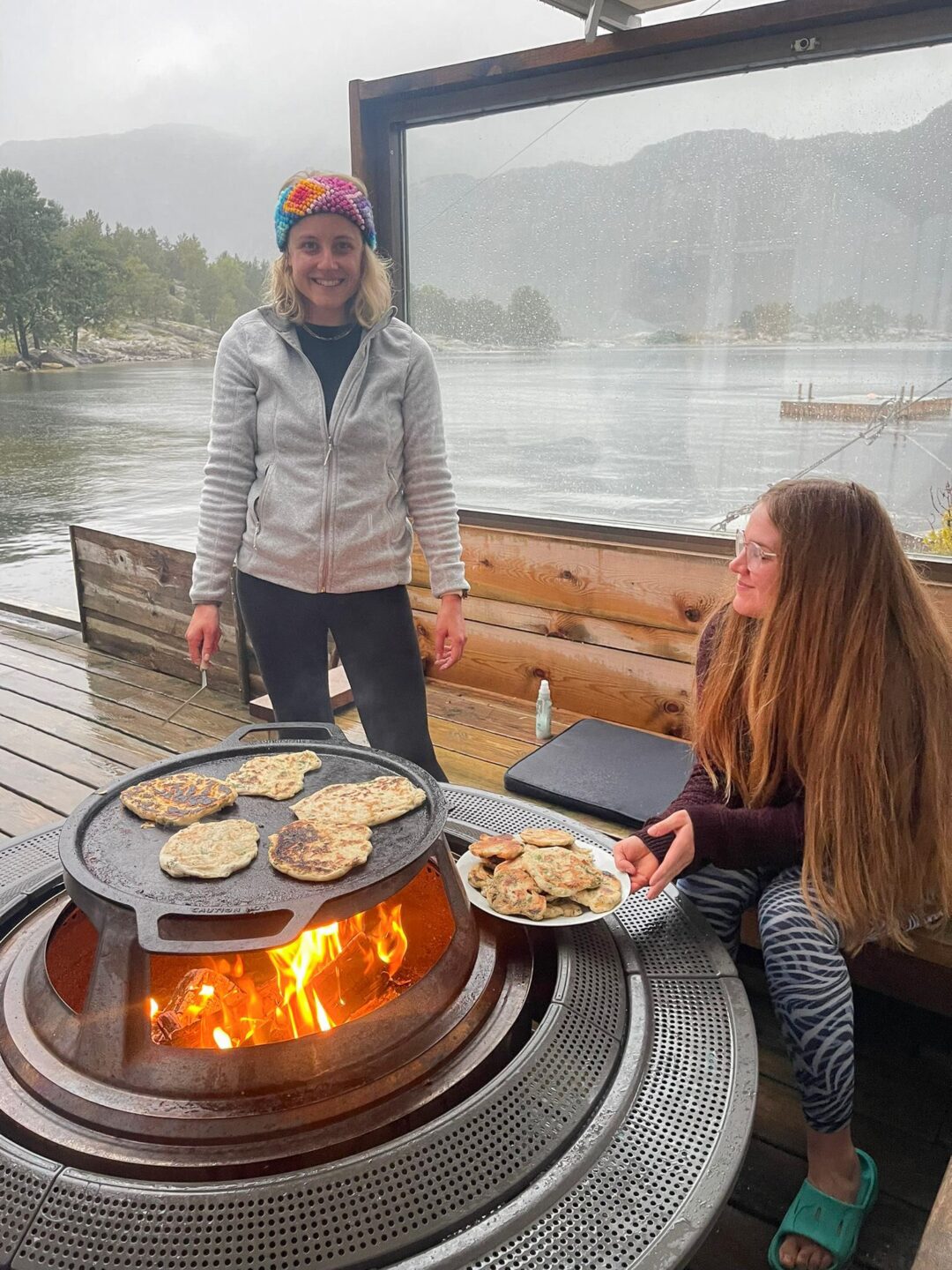 Women cooking by an open fire on a boat by calm fjord water, with mountains in the background