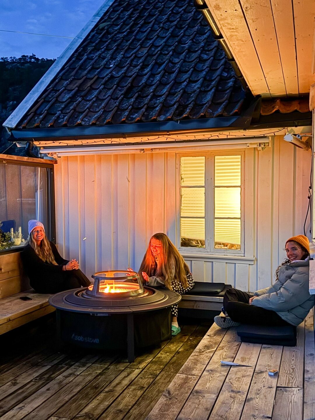 Three women laughing beside an outdoor fire pit on a wooden deck outside a lodge at night