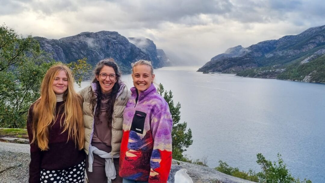 Three women standing on a viewpoint above a Norwegian fjord with misty mountains in the background