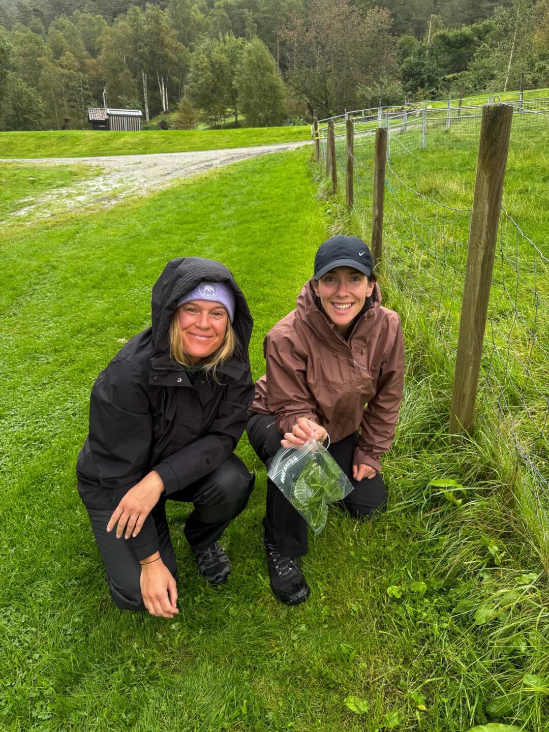 Two women in outdoor waterproofs kneel on a grassy path beside a wooden fence in a forested countryside.