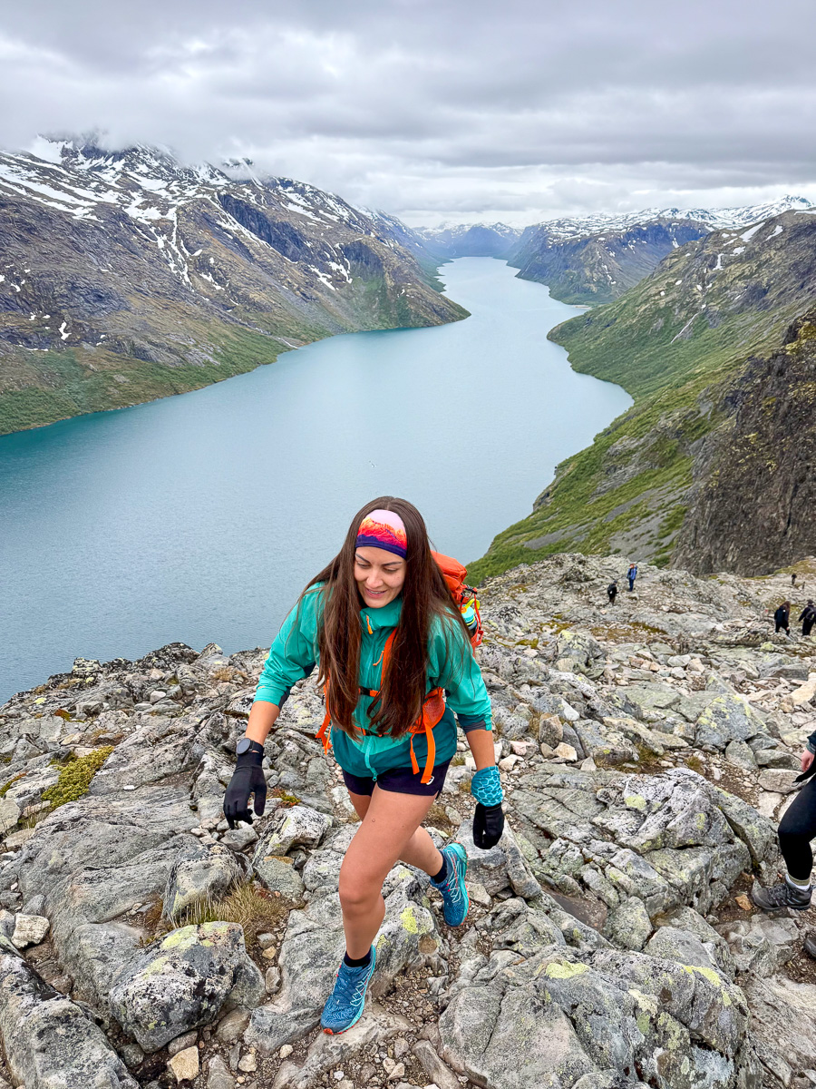 Woman hiking on rocky trail above a fjord in Norway, with snowy mountains and cloudy sky.