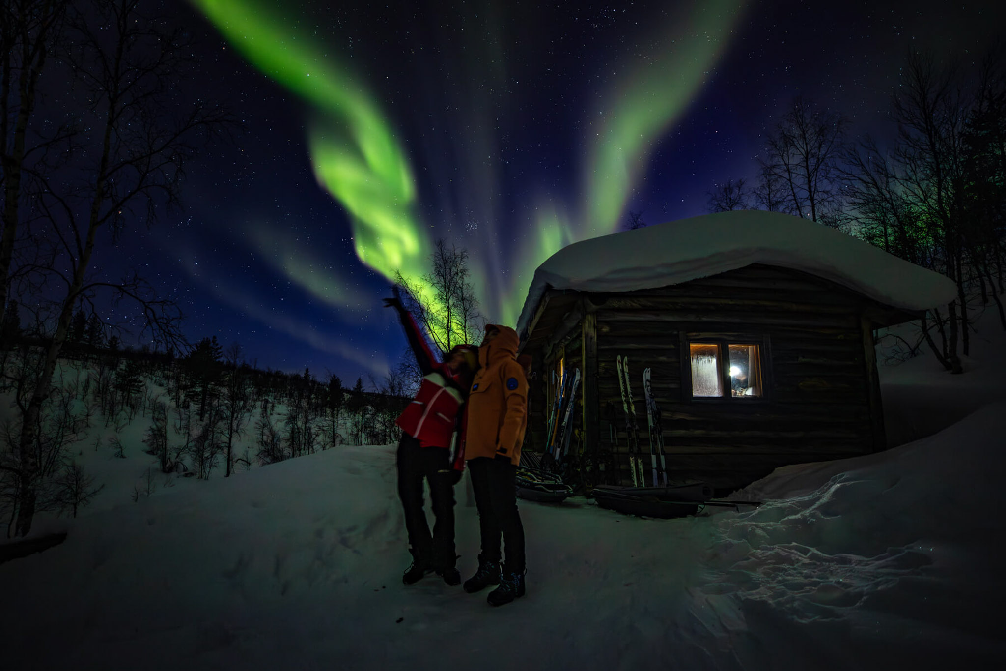 Women stand outside a wooden arctic hut in snowy wilderness under northern lights at night.