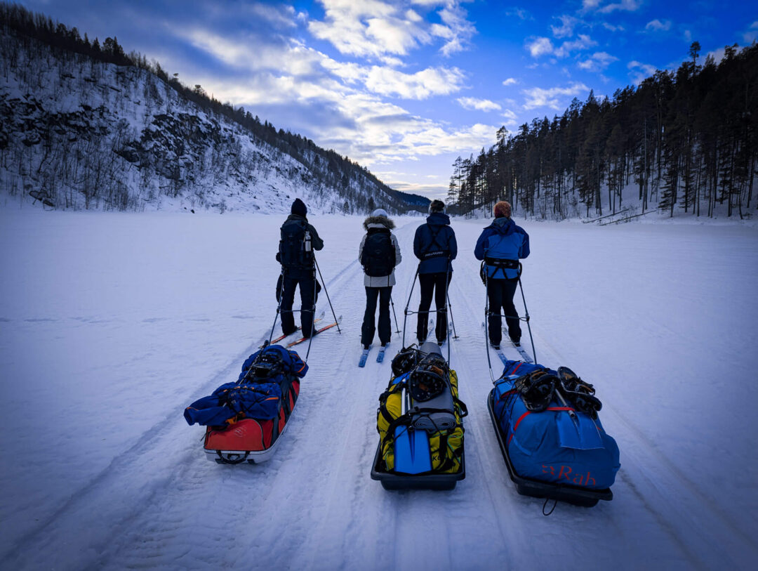 Women cross-country skiing on a snowy trail through a forested valley, with backpacks and sleds ahead