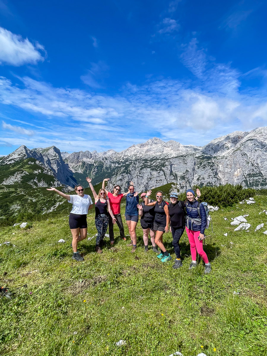 Women hiking across a grassy mountain meadow with rocky peaks under a blue sky