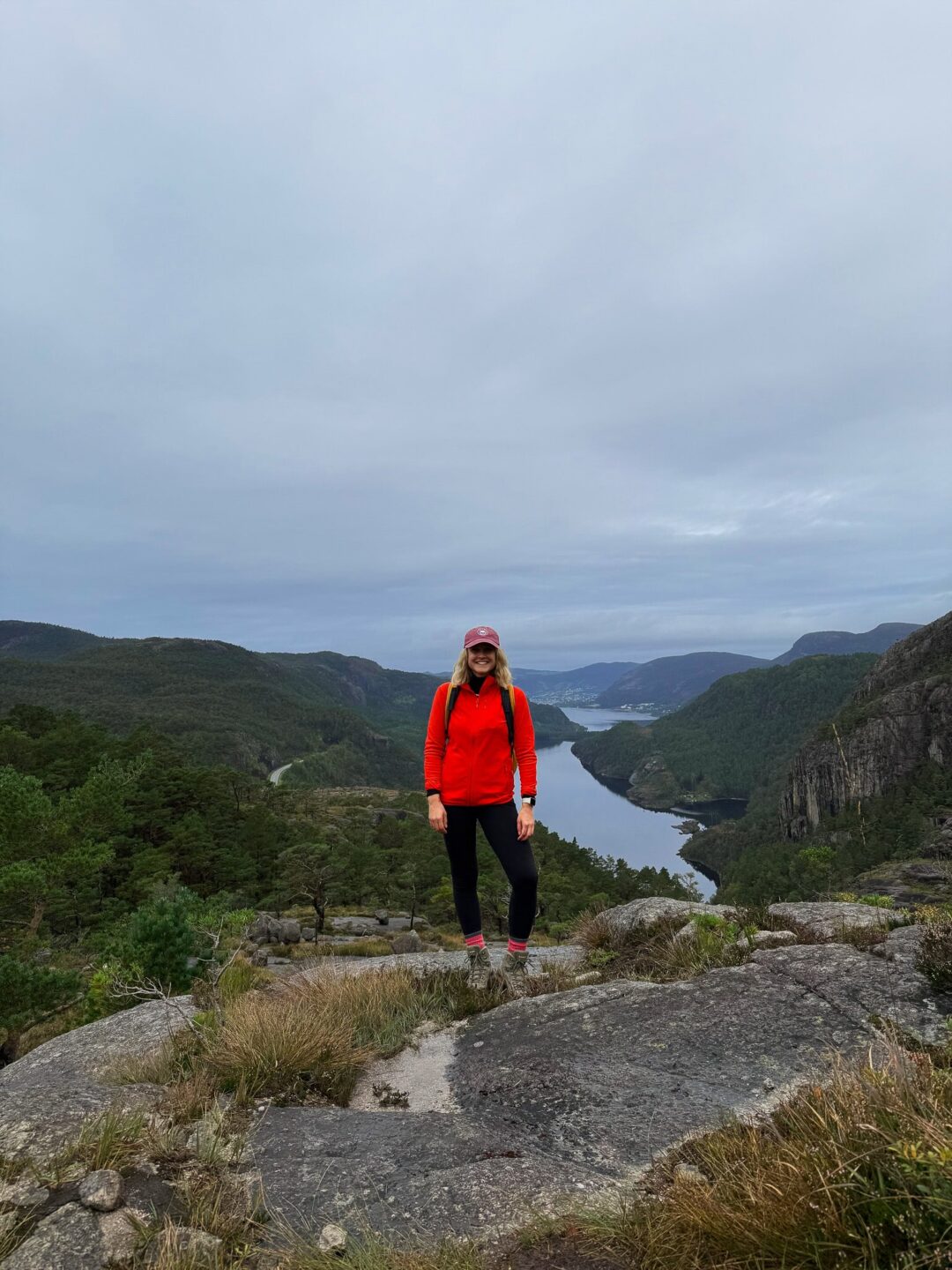 Woman in red jacket and cap standing on a rocky cliff overlooking a fjord valley under cloudy sky