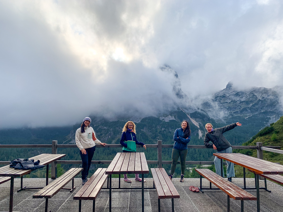 Women hikers at a mountain hut terrace with tables, surrounded by misty alpine peaks