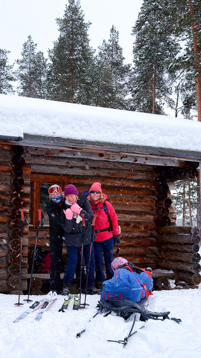 Women in ski gear posing outside a snowy log hut in a pine forest, with skis and backpacks on snow.