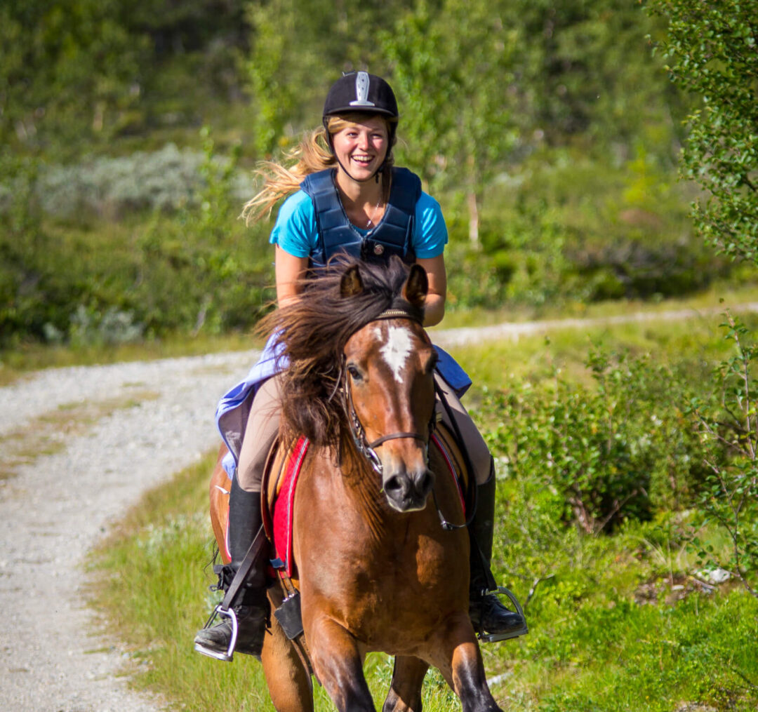 Woman on horseback during an outdoor ride along a woodland trail, wearing a helmet and riding gear.
