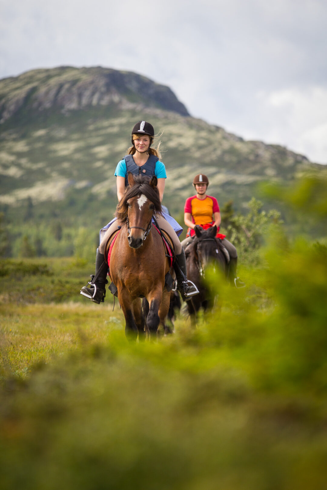 Women riding horses through green valley with mountains in Norway, on an outdoor trail ride.