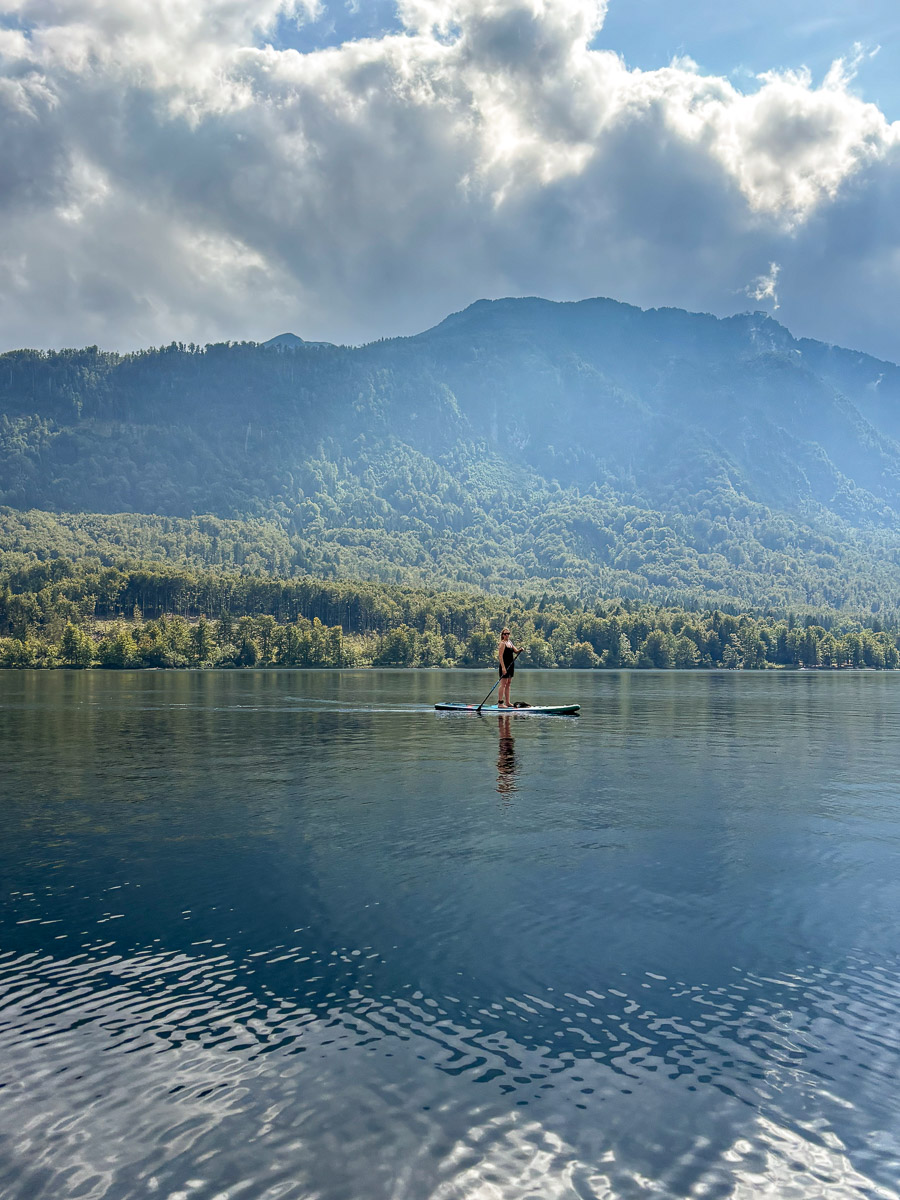 Woman paddleboarding on a mountain lake at sunrise, with forested hills and peaks in the distance.