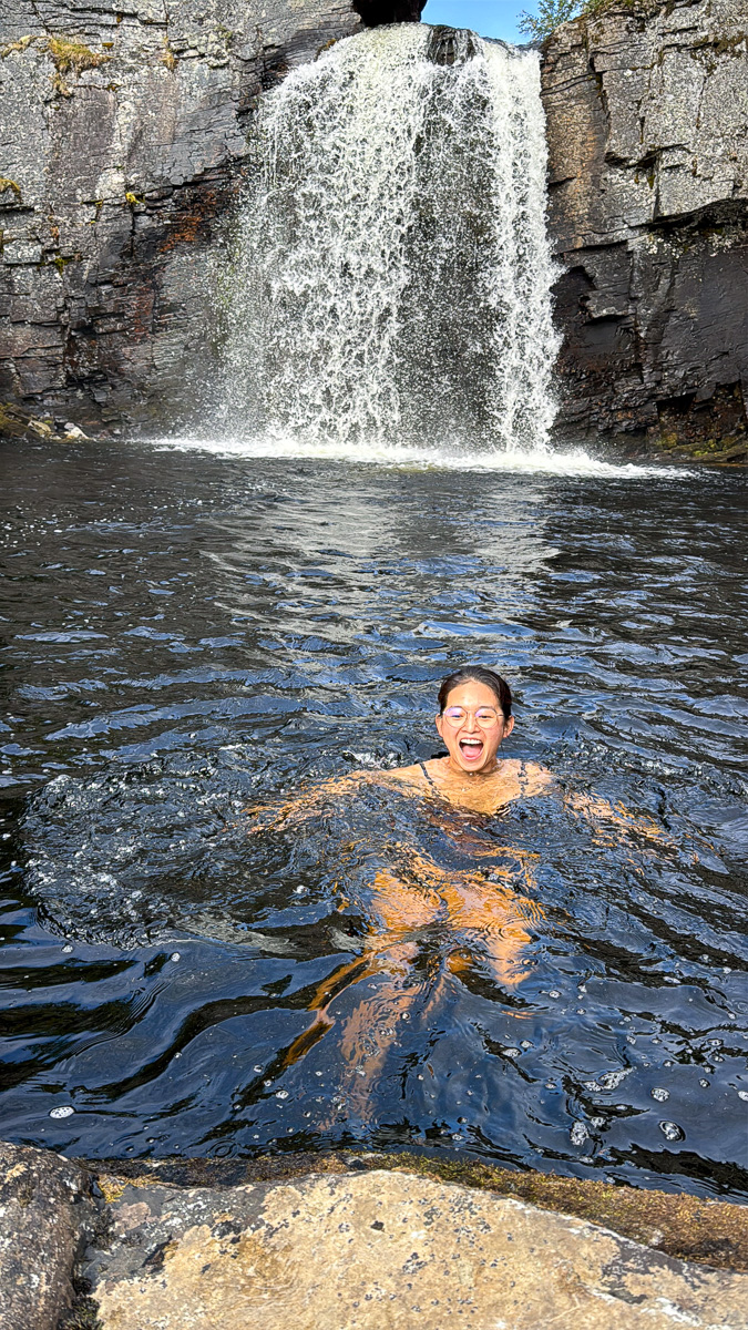 Woman swimming in a river beside a large waterfall in rocky cliffs