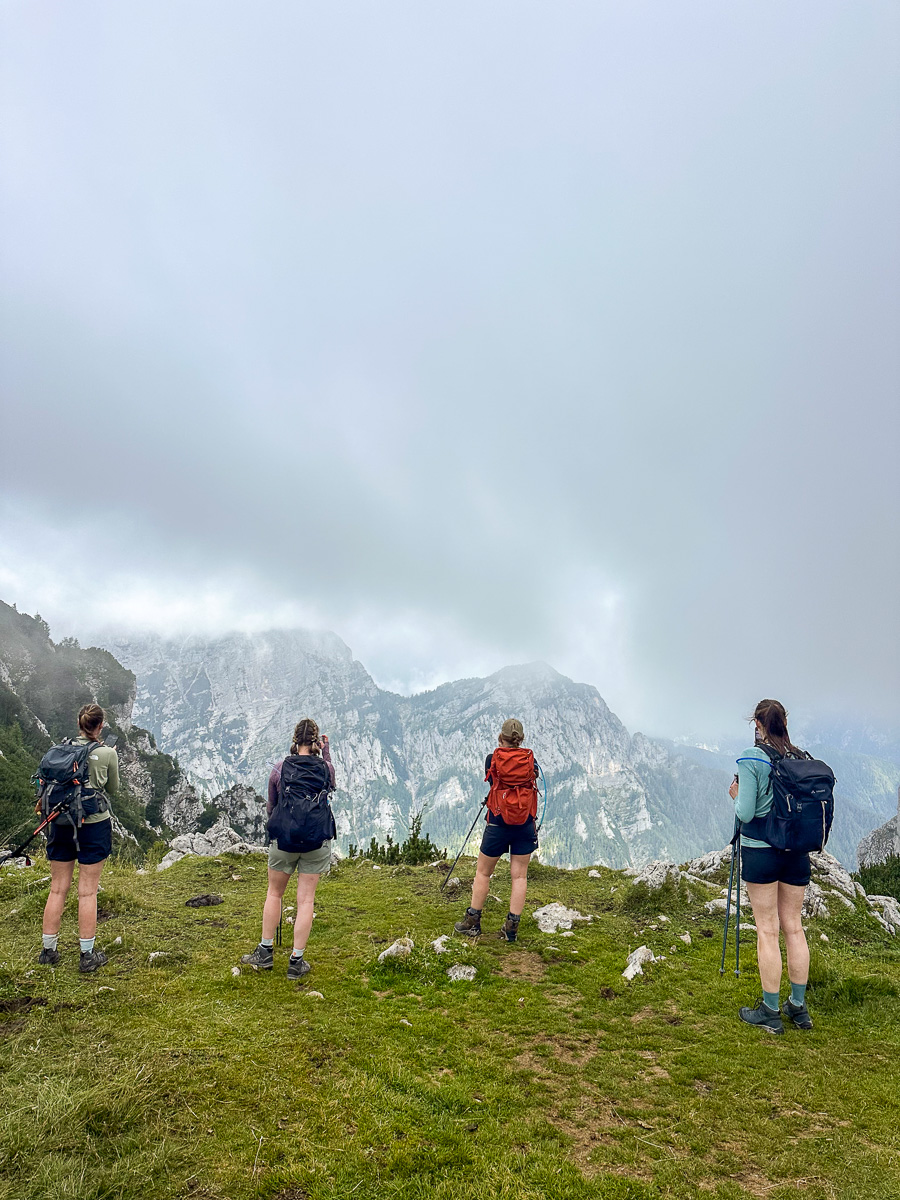 Women hiking on a grassy mountain ridge, with misty alpine peaks in the background