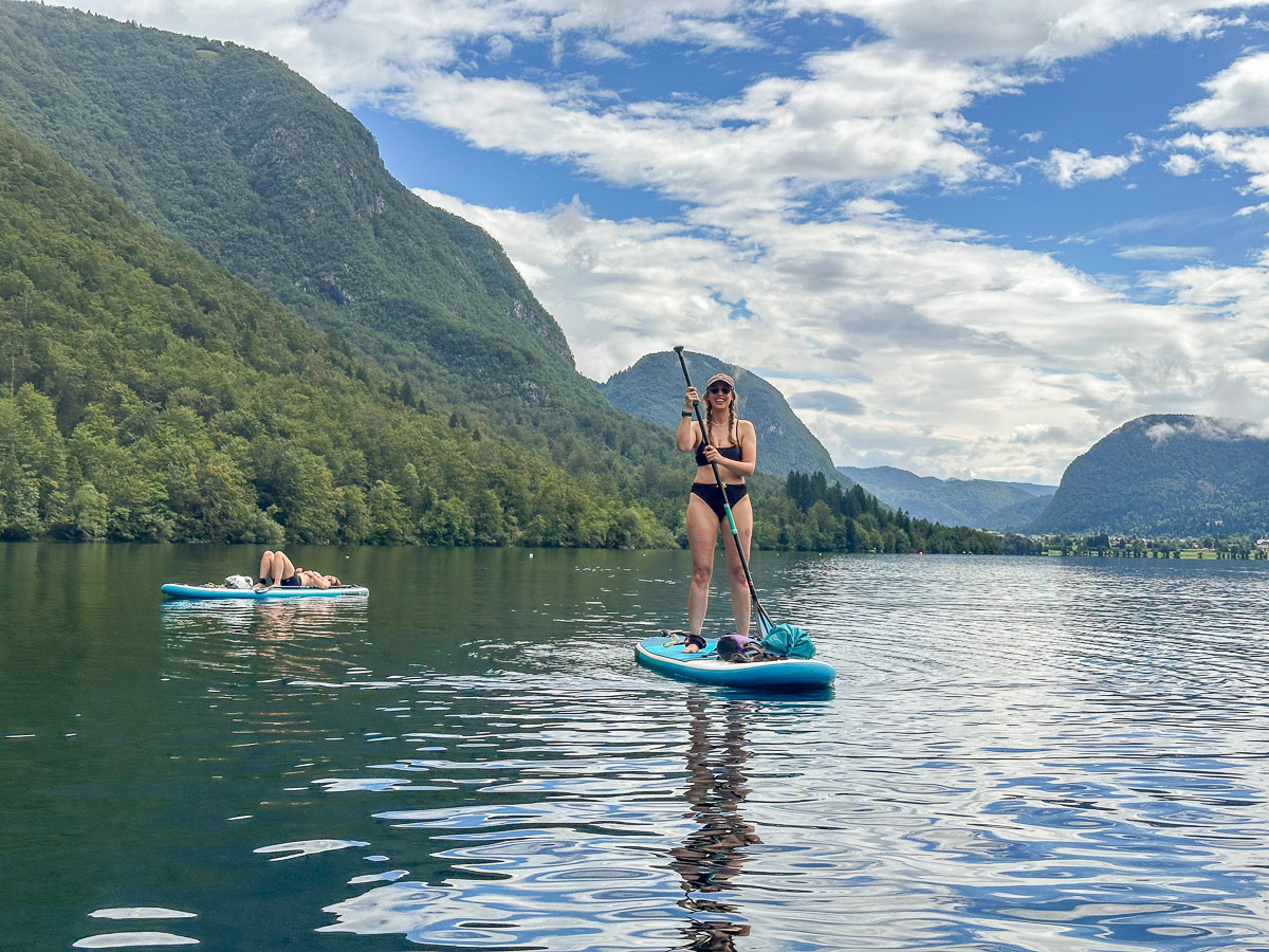 Woman paddleboarding on a mountain lake with forested hills and peaks under a blue sky