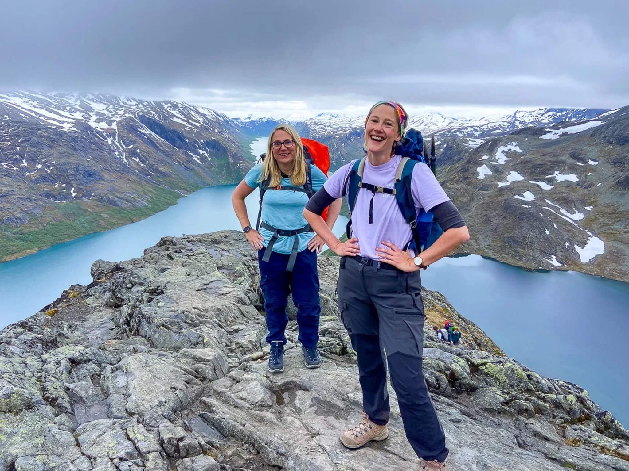 Two women hikers with backpacks on a rocky ridge overlooking a Norwegian fjord and snow-covered mountains