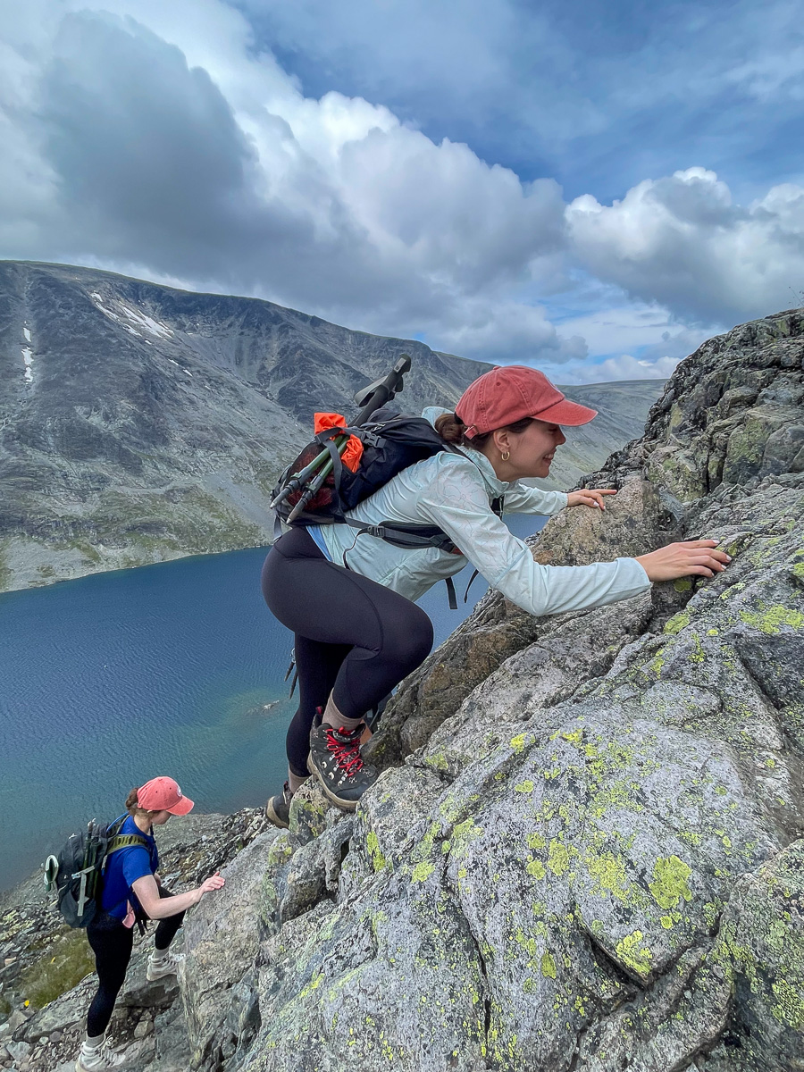 Women hiking on a rocky ridge above a Norwegian fjord, with mountains and clouds overhead