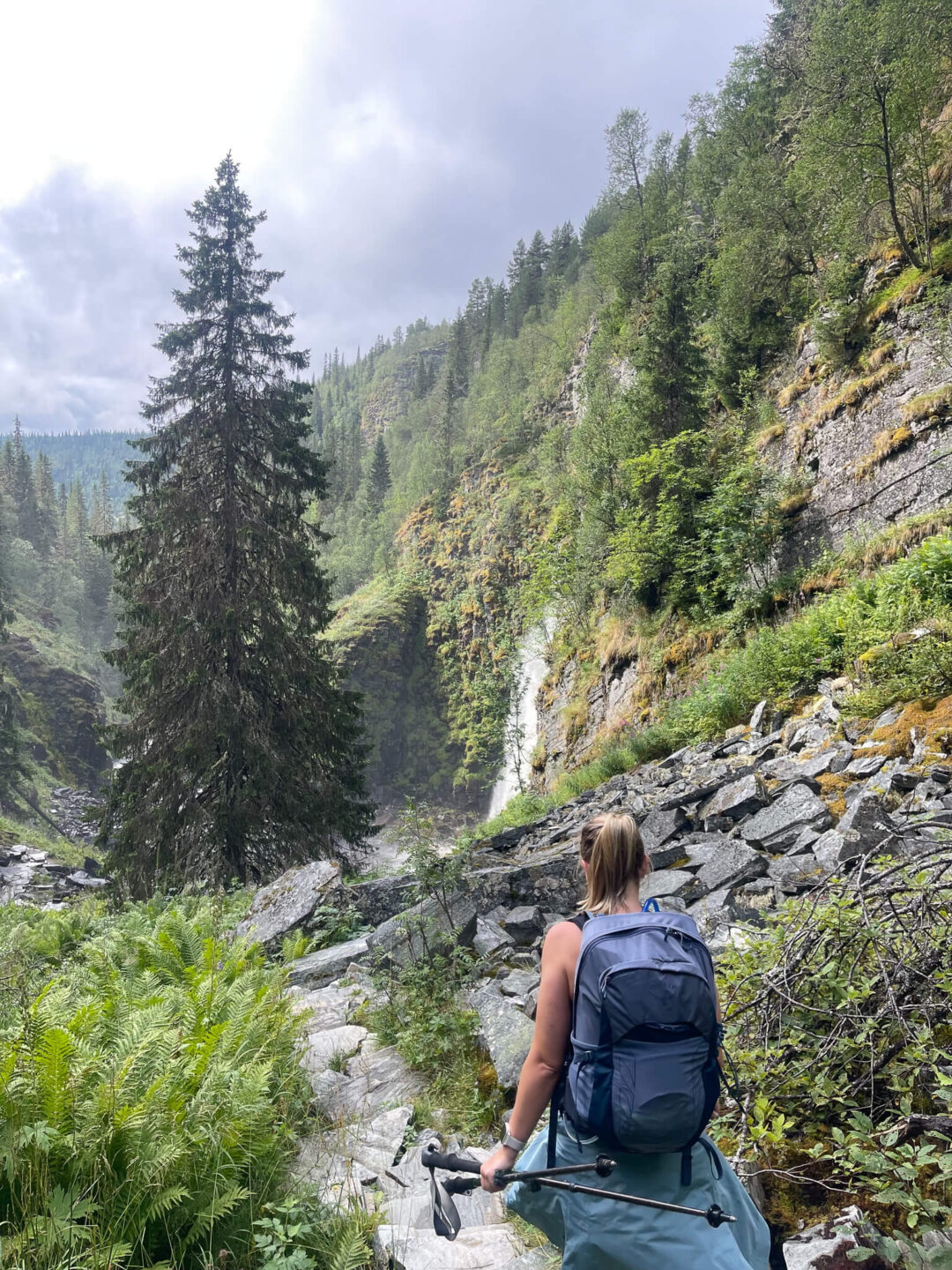 Woman hiking on a rocky trail by a waterfall in Norway’s forest mountains, carrying a backpack