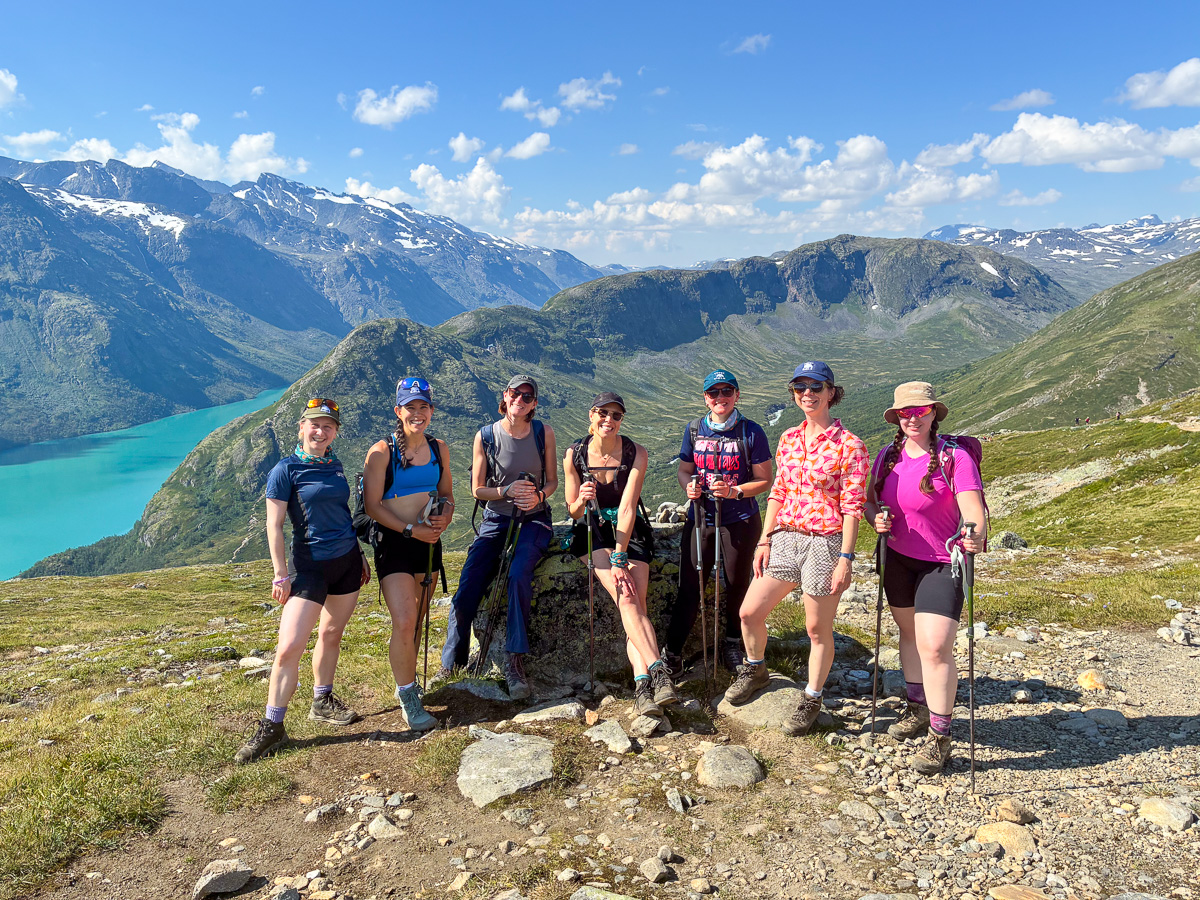 Women hikers posing on a mountain viewpoint above a turquoise lake in Norway, with snowcapped peaks in the background