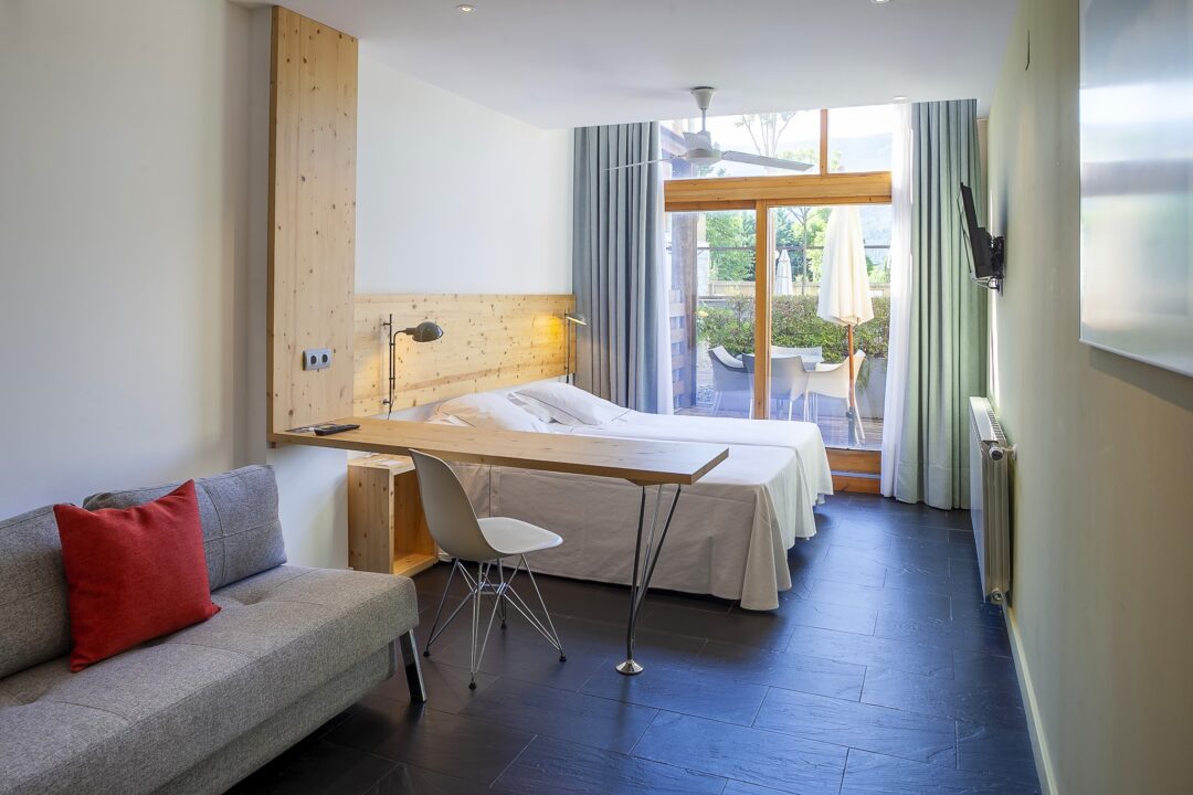 Bright hotel bedroom with double bed, sofa seating, wooden desk and window balcony doors