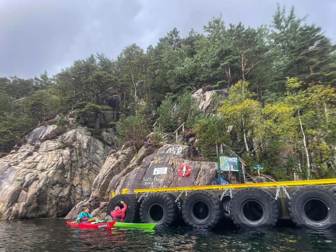 Women kayaking in Norway past a rocky shoreline with tyres used as a water barrier