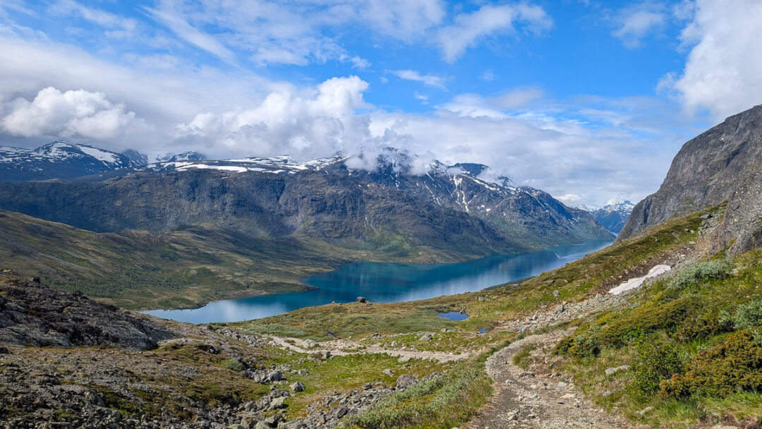 Hiking trail in Norway with a blue mountain lake and snowy peaks under a bright sky